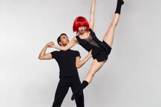 A man wearing all black supports another dancer wearing a red wig as she does a tilt jump.